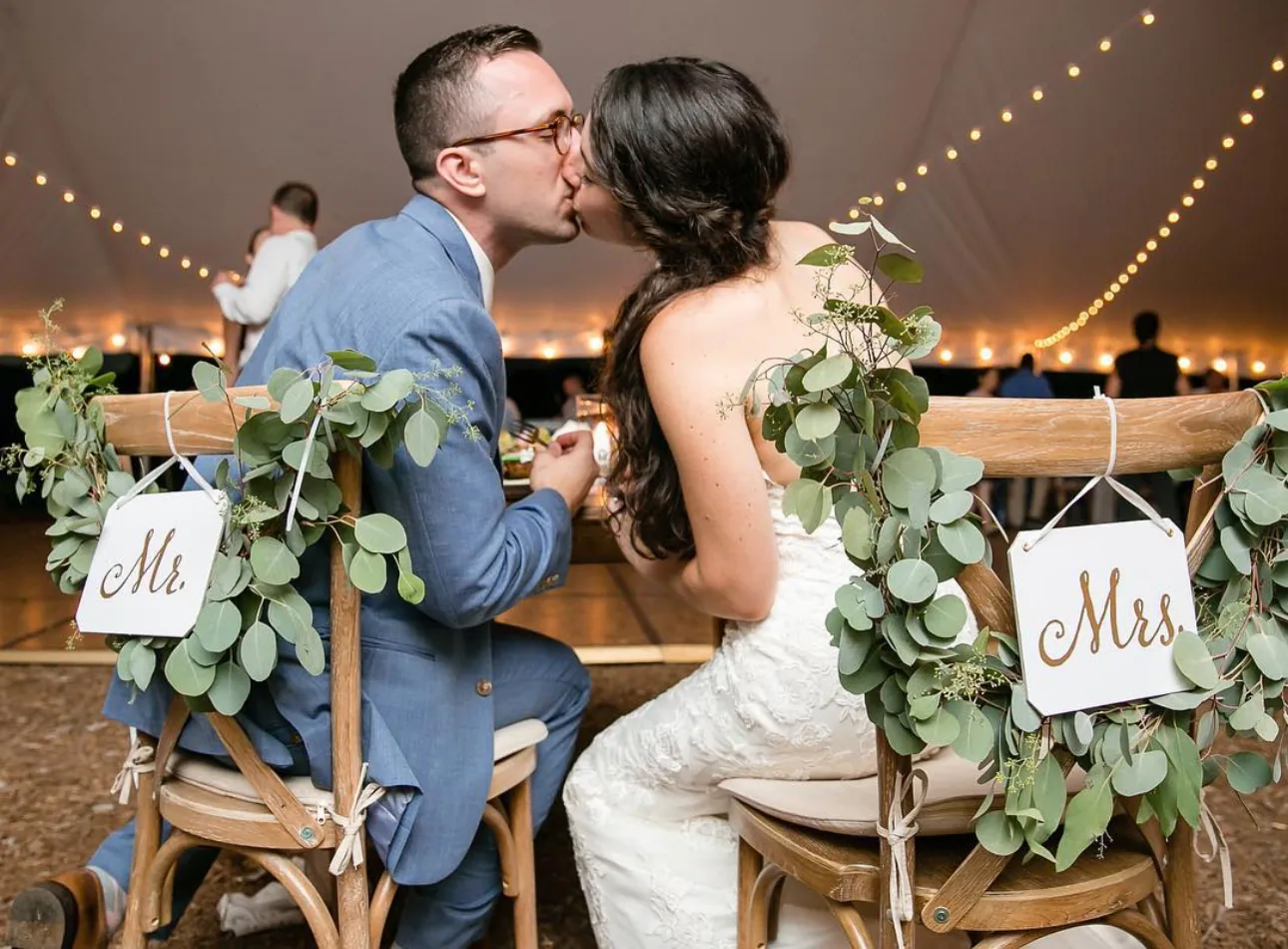 Couple kissing at their sweetheart table, chairs dressed with eucalyptus and Mr and Mrs signs