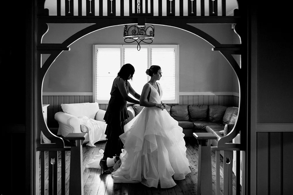 Black and white frame of a bride in a full ruffled gown beside a tall arched window
