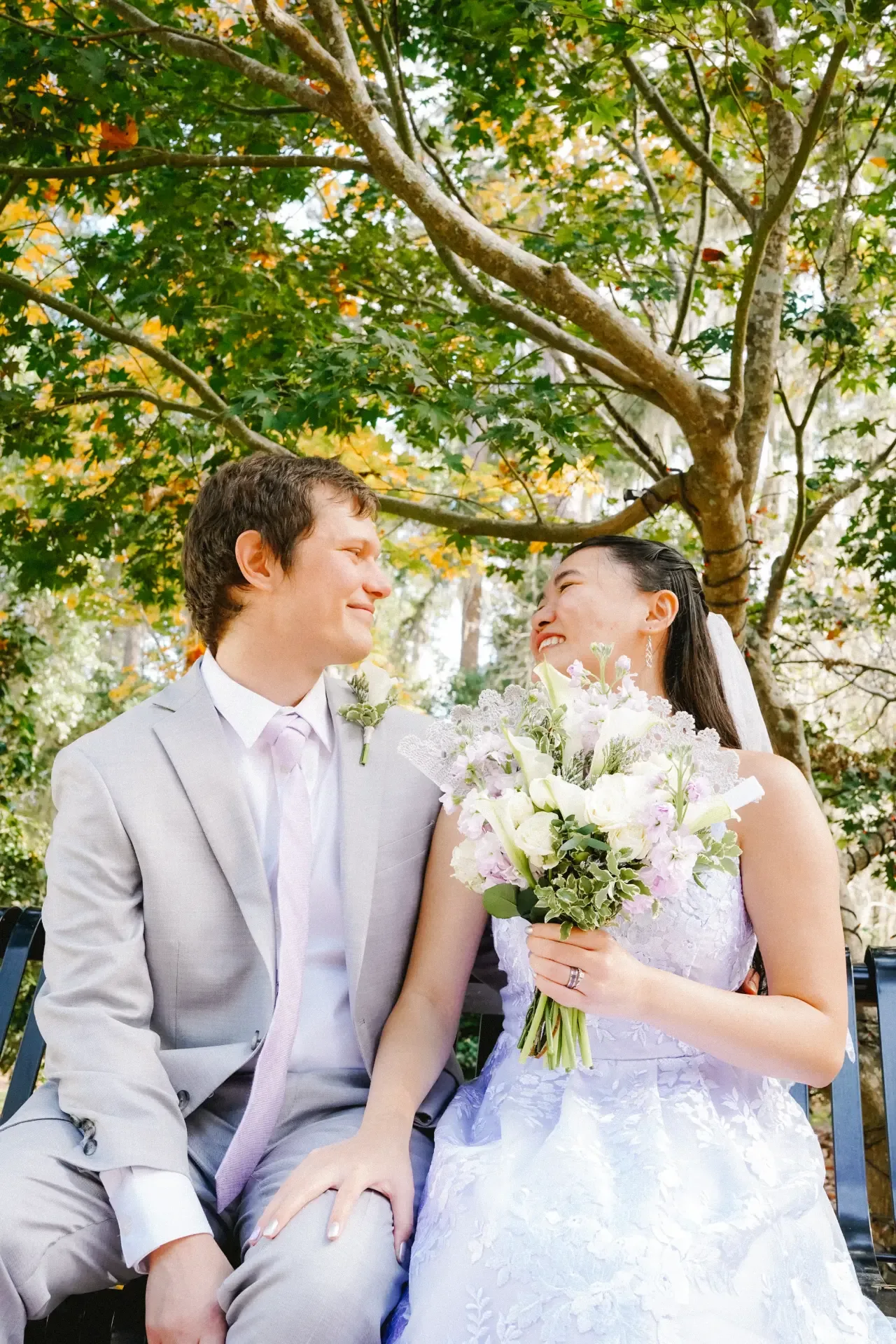 Bride and groom seated on a park bench under autumn trees, laughing together